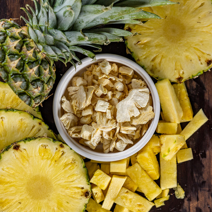 Close-up of Ready Hour freeze-dried pineapple chunks in a white bowl surrounded by slices of fresh pineapple